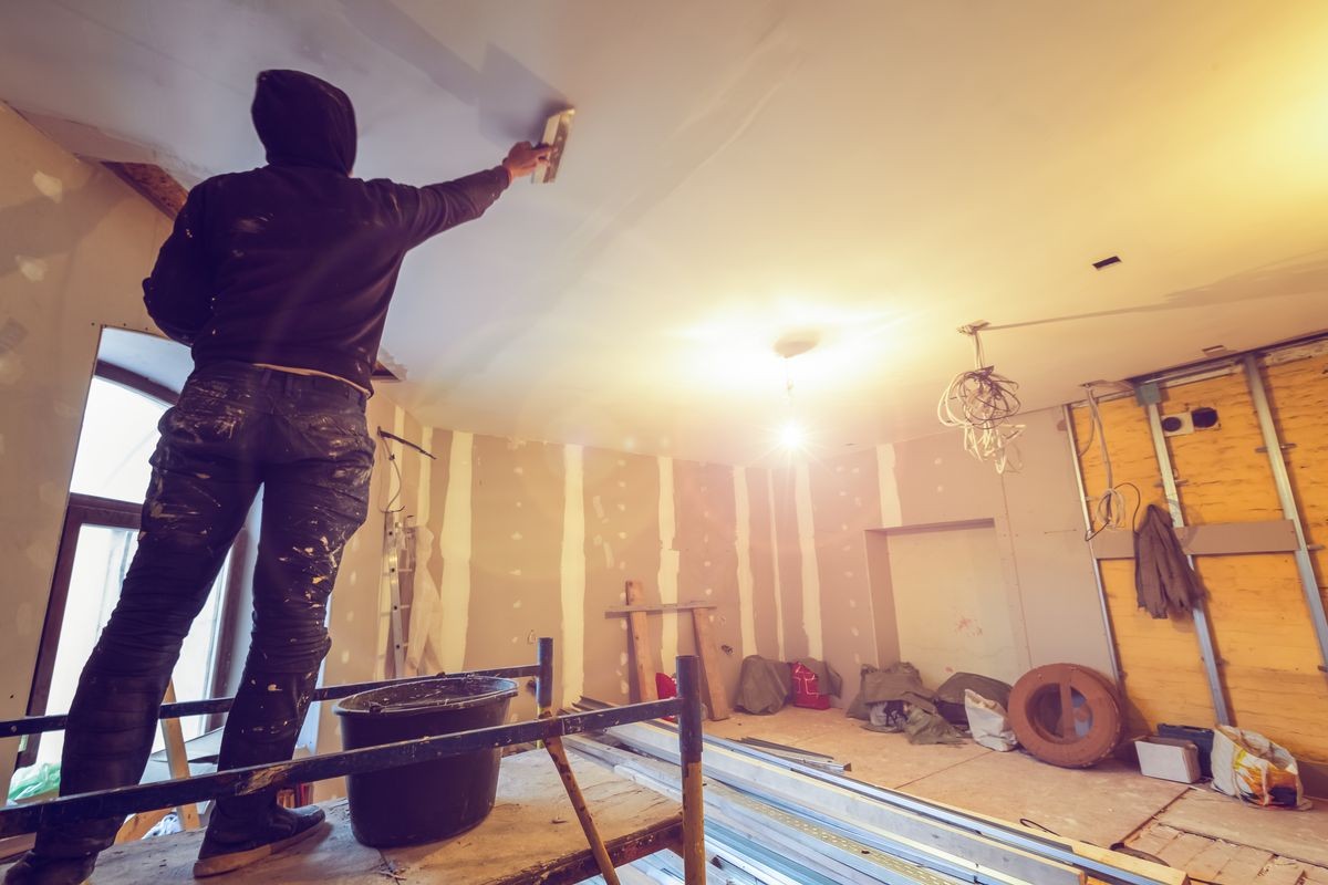 Worker is working with palette-knife for flattening the ceiling from wooden platform in room of apartment is inder construction, remodeling, renovation, overhaul, extension, restoration and